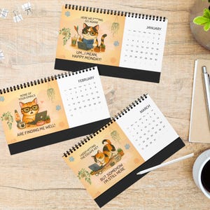 Op de afbeelding: Een spiraalgebonden bureaukalender met drie pagina's voor januari, februari en maart. Elke pagina toont een cartoon kat-illustratie met humoristische tekst, waaronder 'HERE WE F***ING GO AGAIN!', 'NONE OF YOUR EMAILS ARE FINDING ME WELL!' en 'I KEEP HITTING THE ESCAPE KEY BUT SOMEHOW I'M STILL HERE'.