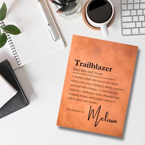 May include: A brown leather-like notebook with the word "Trailblazer" defined as "A fearless spirit who carves new paths where none exist, daring to dream, explore, and lead with courage. A visionary who embraces challenges, turns obstacles into stepping stones, and inspires others to follow their own journey." The text "Also known as: Melissa" is written below the definition.