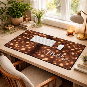 Pode incluir: Um tapete de mesa retangular com um padrão manchado marrom e bege, um teclado branco e um mouse branco. O nome "Maddison" está escrito em branco no tapete. O tapete está em uma mesa de madeira.