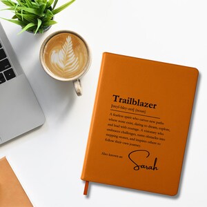 Könnte beinhalten: Ein orangefarbenes Notizbuch mit dem Wort "Trailblazer" und seiner Definition auf dem Einband. Das Notizbuch trägt auch den Namen "Sarah" auf dem Einband. Eine Tasse Kaffee und eine kleine Pflanze sind ebenfalls im Bild.