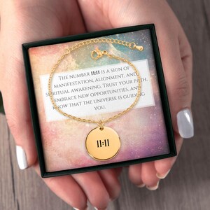 May include: A gold-colored bracelet with a circular pendant featuring the number 11:11. The bracelet is presented in a black box with a printed message about manifestation and spiritual awakening. The background has a colorful, cosmic design.