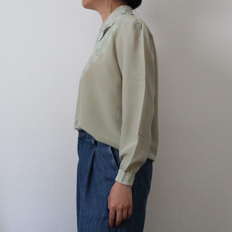May include: A light green long-sleeve blouse with a delicate, embroidered collar. The blouse is paired with blue denim jeans. The sleeves have buttoned cuffs. The blouse is made of a soft, flowing fabric.