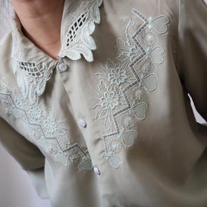 May include: A light green blouse with intricate floral and geometric embroidery in a lighter green shade. The blouse features a scalloped collar and button-down front, offering a vintage aesthetic. The embroidery includes flowers, hearts, and geometric patterns.