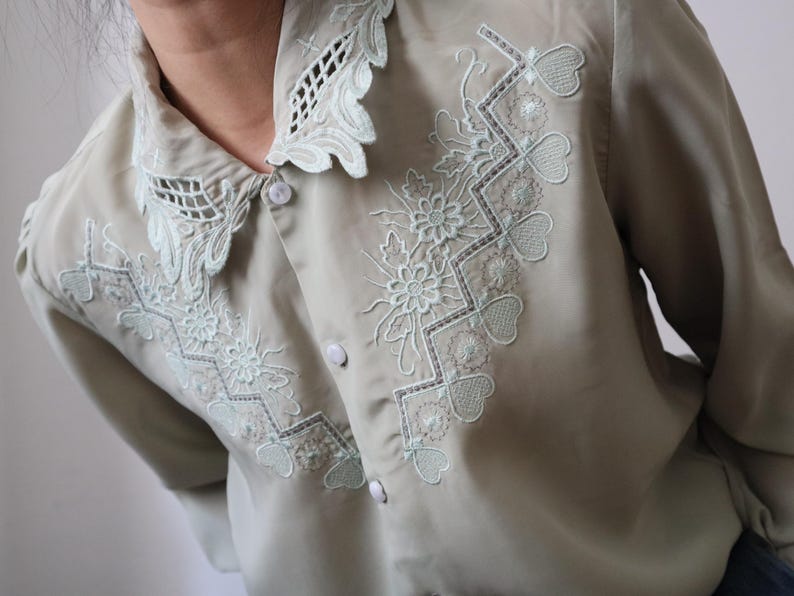 May include: A light green blouse with intricate floral and geometric embroidery in a lighter green shade. The blouse features a scalloped collar and button-down front, offering a vintage aesthetic. The embroidery includes flowers, hearts, and geometric patterns.