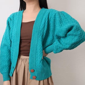 Peut inclure: Un cardigan turquoise avec un motif texturé et un col en V. Le cardigan a des manches longues et deux boutons marron. Le modèle porte un haut marron et une jupe beige.