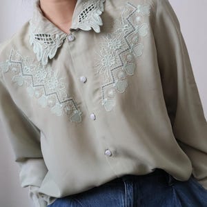 May include: A light green button-down shirt with intricate floral and heart-shaped embroidery in a lighter green shade. The shirt features a delicate lace collar and long sleeves, offering a vintage aesthetic.