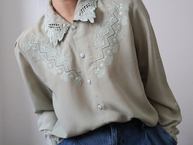 May include: A light green button-down shirt with intricate floral and heart-shaped embroidery in a lighter green shade. The shirt features a delicate lace collar and long sleeves, offering a vintage aesthetic.