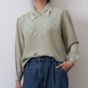 May include: A sage green button-down blouse with a delicate, embroidered collar and yoke. The long-sleeved shirt features a detailed floral design in a lighter green shade. The blouse is paired with blue denim jeans.
