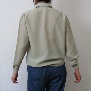 May include: A light green long-sleeve blouse with a ruffled collar and gathered shoulders. The blouse is paired with blue denim jeans. The back view of the blouse shows the details of the design and fit.