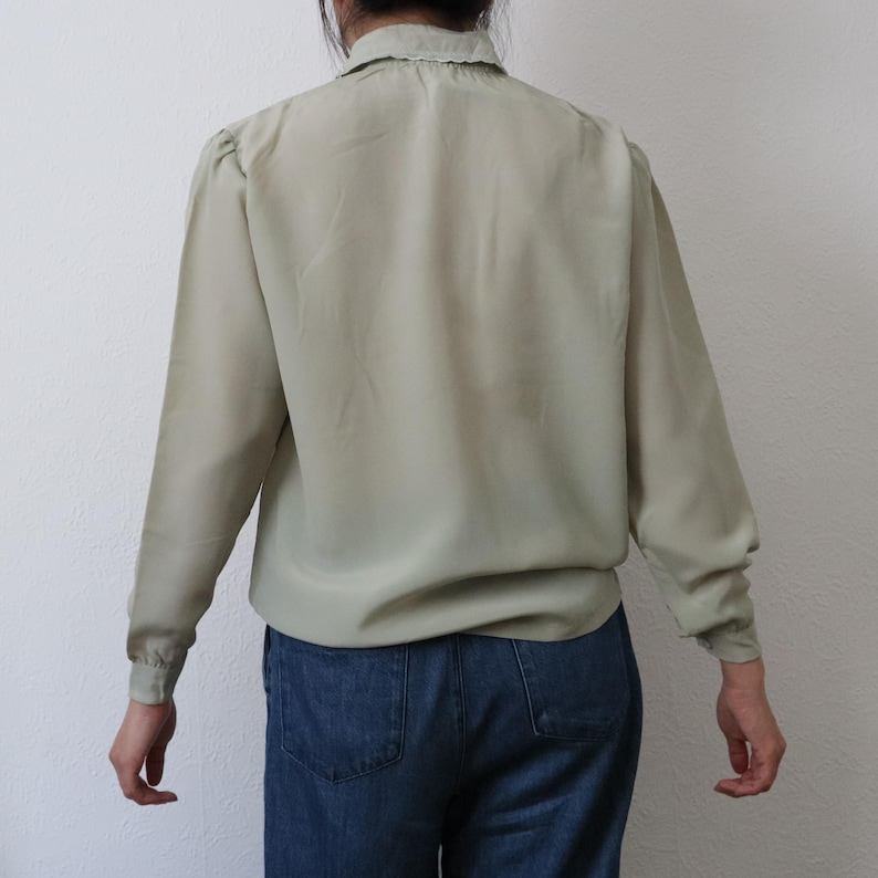 May include: A light green long-sleeve blouse with a ruffled collar and gathered shoulders. The blouse is paired with blue denim jeans. The back view of the blouse shows the details of the design and fit.