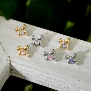 May include: Six bow-shaped stud earrings in gold and silver. Each earring has a central gemstone in clear, pink, blue, or purple. The earrings are displayed on a white surface with a blurred green background.