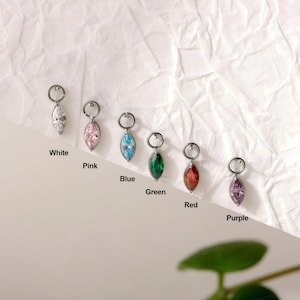 Könnte beinhalten: Sechs silberfarbene Creolen, jede mit einem marquise-geschliffenen Edelstein in Weiß, Pink, Blau, Grün, Rot und Violett. Jeder Ohrring ist unterhalb mit seinem Farbnamen beschriftet und in einer diagonalen Linie auf Weiß angeordnet.