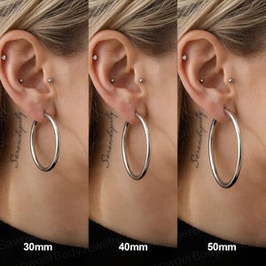 May include: Three silver-toned hoop earrings shown in a side-by-side comparison. The earrings are labelled 30mm, 40mm, and 50mm, indicating their respective diameters. Each hoop is a simple, smooth, circular metal band worn in an earlobe piercing.