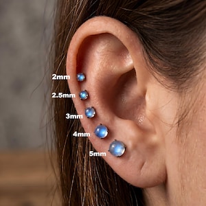 May include: Close-up of an ear with multiple sizes of moonstone stud earrings. The sizes are 2mm, 2.5mm, 3mm, 4mm, and 5mm. The earrings have a silver setting and a blue-white iridescent stone.