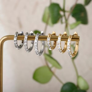 Op de afbeelding: Een collectie van zes oorringen gerangschikt op een goudkleurige displaystang. Er zijn drie zilverkleurige en drie goudkleurige ringen te zien, elk bezet met een rij heldere, sprankelende steentjes langs de buitenrand van de oorring.