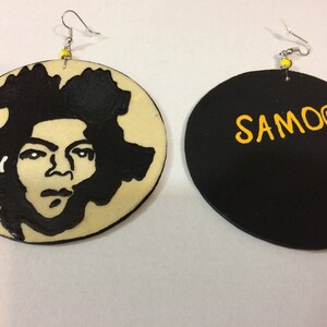 Op de afbeelding: Een paar ronde houten oorbellen. E&eacute;n oorbel heeft een zwart-wit silhouet van een menselijk gezicht. De andere oorbel is zwart met het woord "SAMOO" in geel.