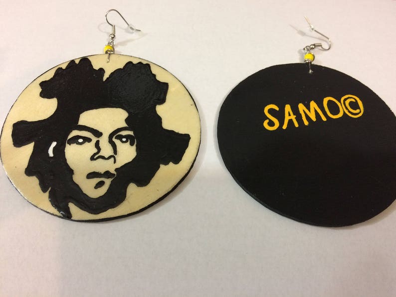 Op de afbeelding: Een paar ronde houten oorbellen. E&eacute;n oorbel heeft een zwart-wit silhouet van een menselijk gezicht. De andere oorbel is zwart met het woord "SAMOO" in geel.