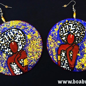May include: A pair of round, hand-painted wooden earrings. Each earring features a stylized figure with a red body and a white and black afro against a blue background with gold flecks. The earrings have silver-colored hooks.