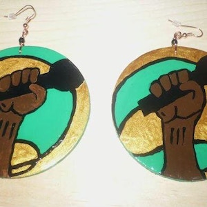 May include: A pair of round wooden earrings with a green, gold, and black design. The design features a raised fist holding a microphone.