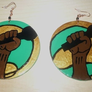 May include: Hand-painted wooden earrings featuring a brown fist holding a black microphone against a green and gold background. The earrings have copper-colored ear wires and black beads.
