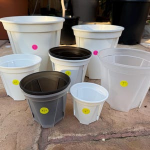 May include: A collection of white and dark gray plastic plant pots of various sizes. Some pots have yellow price tags with inch measurements like 4", 4.7", 5.5", and 7". The pots are designed for planting flowers or other plants.