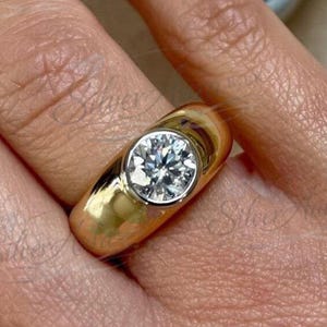 May include: A gold ring with a large, round, clear gemstone set in a bezel. The ring is on a finger, and the gold band is thick and polished. The gemstone sparkles, reflecting light.