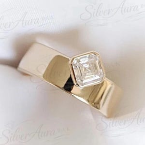 Esta versión del anillo de compromiso descentrado está hecha con una moissanita talla Asscher de 1,25 quilates engastada en bisel, y el diamante engastado en una esquina de la banda ancha.