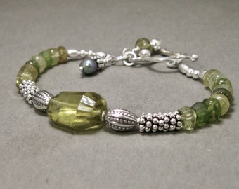 Puede incluir: Un brazalete verde y plateado con una gran piedra preciosa verde claro facetada. El brazalete tiene un cierre de palanca y un pequeño amuleto de perla gris oscuro.