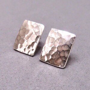 May include: A pair of silver-toned square stud earrings. Each earring has a hammered texture, creating a dimpled surface. The earrings are displayed on a gray surface.