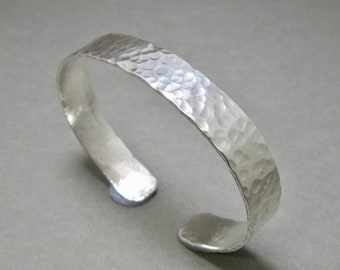 May include: A silver cuff bracelet with a hammered texture. The open-ended bracelet has a slightly curved shape and a textured surface. The metal reflects light, creating highlights and shadows. The bracelet is a simple, elegant piece of jewelry.