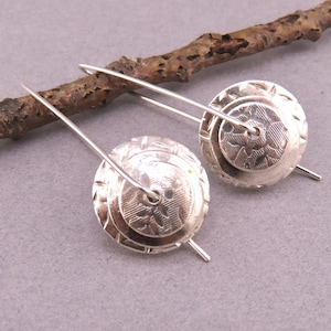 May include: A pair of silver earrings with a circular design. Each earring features a layered, embossed pattern with floral and geometric details. The earrings are suspended from long, thin silver wires, and rest on a gray surface.