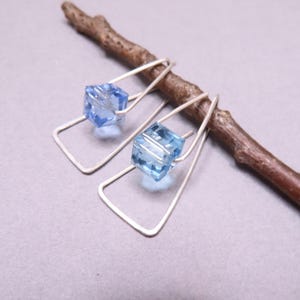 May include: A pair of earrings featuring a silver-toned wire frame in a trapezoidal shape. Each earring holds a single, faceted, light blue cube-shaped bead suspended within the wire structure. The earrings are displayed resting on a brown twig.