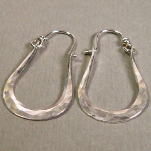 May include: A pair of silver teardrop-shaped earrings. The earrings have a hammered texture and a hook closure. The earrings are displayed against a neutral gray background. The earrings are a jewelry item.