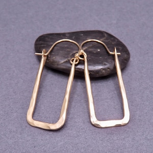 May include: A pair of gold-colored, rectangular hoop earrings with a hammered texture. The earrings are displayed on a dark, oval-shaped stone against a gray background. The earrings have a simple, modern design.