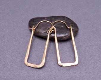 Könnte beinhalten: Ein Paar goldfarbene, rechteckige Creolen mit gehämmerten Texturen. Die Ohrringe werden auf einem dunklen, oval geformten Stein vor grauem Hintergrund präsentiert. Die Ohrringe haben ein schlichtes, modernes Design.