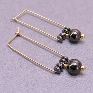 May include: A pair of gold-colored, rectangular hoop earrings. Each earring features a cluster of small, dark gray beads, a larger dark gray sphere, and a small gold-colored bead. The earrings are displayed on a gray surface.