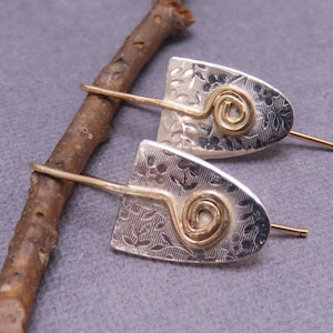 May include: A pair of silver earrings with a floral pattern and gold accents. Each earring features a half-moon shape with a spiral design in the center. The earrings are displayed on a gray surface with a small branch.