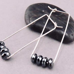 May include: Silver-coloured, rectangular hoop earrings. Each earring has five faceted, dark grey beads. The earrings are displayed on a dark grey, smooth stone against a light grey background.