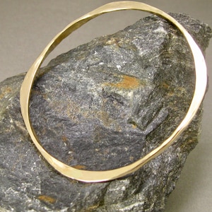 May include: A gold-colored, hammered metal bangle bracelet. The bracelet has an irregular, organic shape and is displayed on a dark gray, textured rock. The bangle is a simple, elegant piece of jewelry.