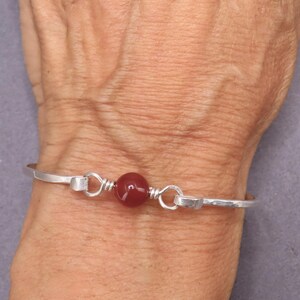 May include: A silver bracelet with a central, round, deep red bead. The bracelet has a simple design with a hammered texture and a hook closure. The bead is wire-wrapped and adds a pop of color.