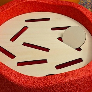 May include: A close-up of a red, textured, circular object with a light-colored interior. The interior has several red rectangular cutouts and a circular piece. The object is likely a toy or puzzle.