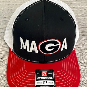 May include: A black, red, and white trucker cap featuring the letters "MAGA" in white, and a red and black "G" logo. The cap has a red peak, a black front panel, and a white mesh back. It is adjustable and has a red button on top.