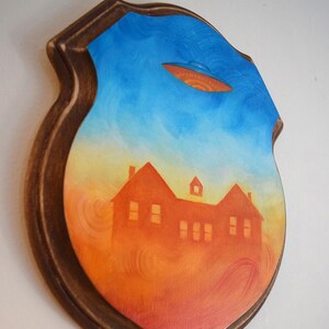 May include: A wooden plaque with a painted scene of a house under a sunset sky, with a UFO hovering above. The artwork is framed in a dark brown wooden border. The artist's signature is in the bottom right corner.