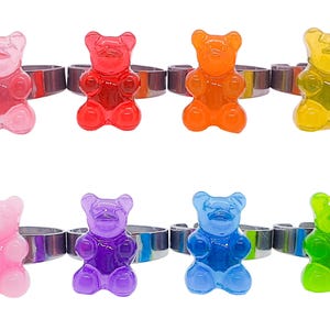 Puede incluir: Conjunto de diez anillos de osito de goma en colores surtidos, incluyendo rosa, rojo, naranja, amarillo, morado, azul y verde. Cada anillo presenta un diseño de osito de goma.