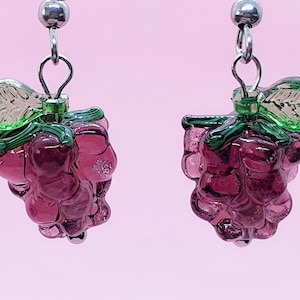 Op de afbeelding: Oorbellen in de vorm van druiven. Elke oorbel heeft een cluster van doorschijnende paarse kralen, groene bladeren en zilverkleurige metalen fournituren. De oorbellen hangen aan een zilverkleurige bal.