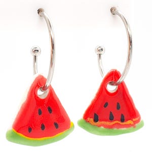 May include: A pair of silver hoop earrings featuring watermelon slice pendants. The pendants are red with black seeds and green rinds. The earrings are displayed against a white backdrop.