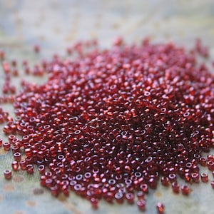 May include: A large pile of small, shiny, red glass beads. The beads are scattered on a light-colored fabric surface.
