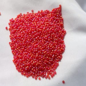 May include: A large pile of small, round, red and orange iridescent glass beads. The beads are scattered on a white surface.