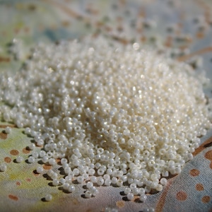 May include: A pile of small, iridescent white beads. The beads are scattered on a patterned fabric with a mix of colors, including green, blue, and brown. The beads have a shiny, pearlescent quality.