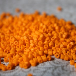 May include: A large pile of small, bright orange plastic beads. The beads are round with a hole in the center. They are scattered on a light gray fabric surface.